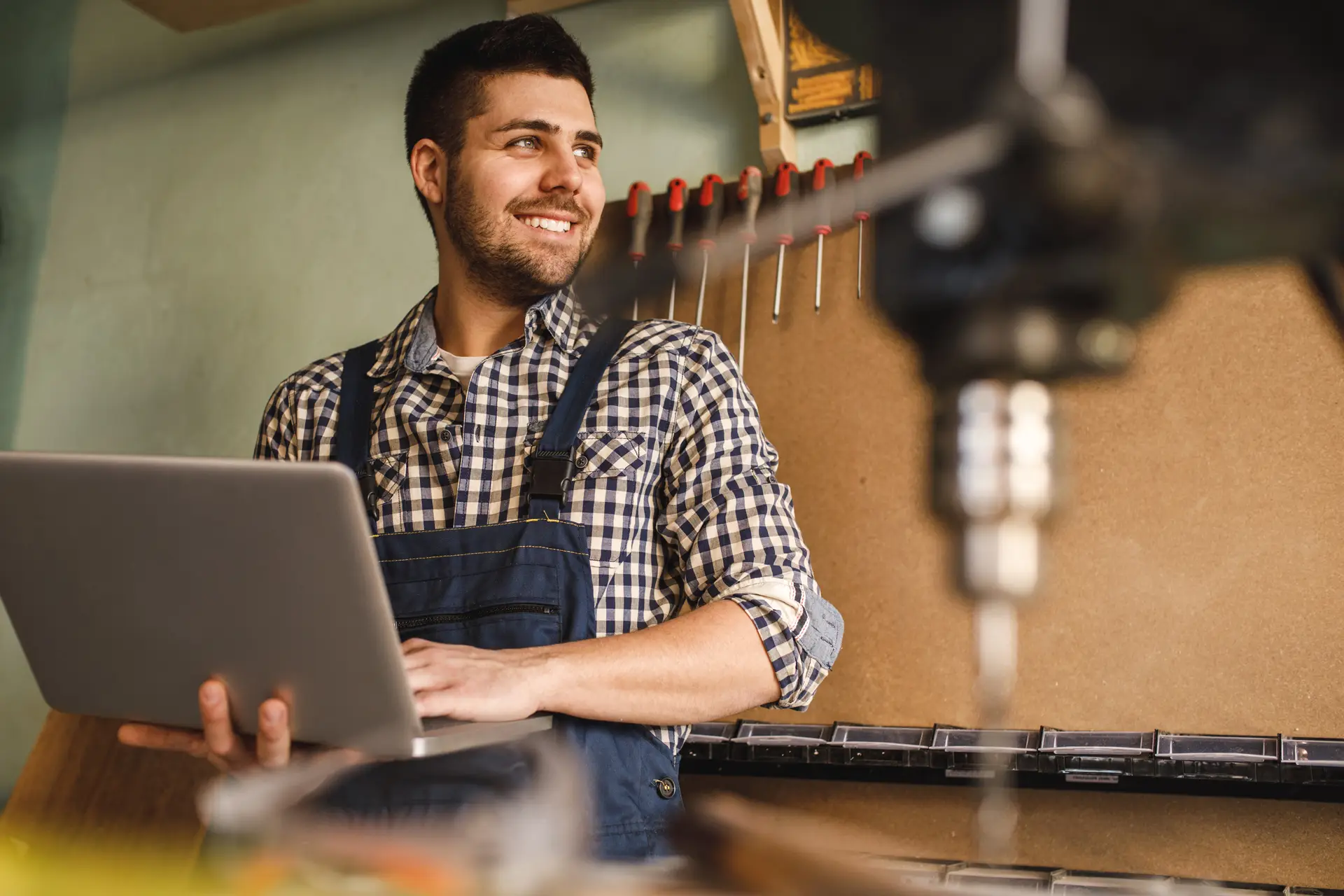 A smiling person in a workshop holds a laptop while standing in front of a tool wall. They are wearing a plaid shirt and dark work overalls.
