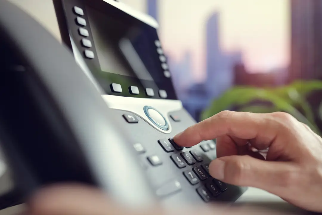 Hand pressing buttons on a desk phone keypad in an office setting.