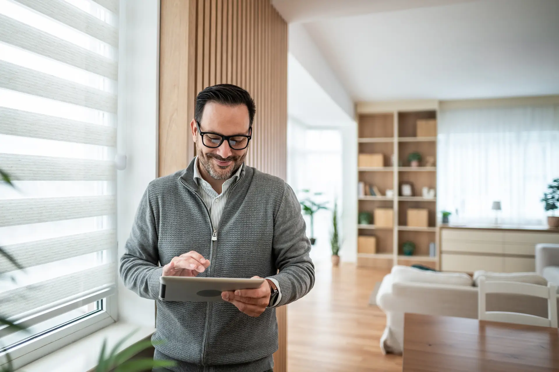 A man wearing glasses and a gray zip-up sweater stands in a bright, modern home, smiling as he uses a tablet. Large windows provide natural light, and a living area with shelving, plants, and light-colored furnishings is visible in the background.
