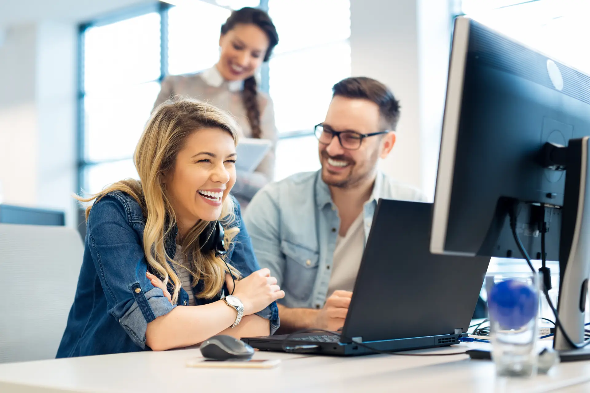 Three coworkers smiling and collaborating at a desk while looking at a computer screen in a modern office.