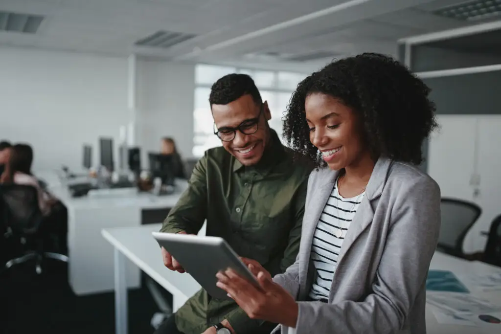 Two coworkers smiling while reviewing content on a tablet in a modern office.
