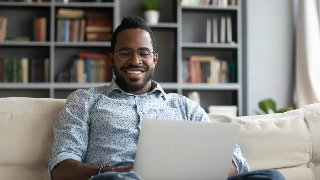 A smiling man wearing glasses sits comfortably on a sofa, using a laptop in a bright living room. A bookshelf filled with books and decorative items is visible in the background, creating a relaxed and inviting home environment.