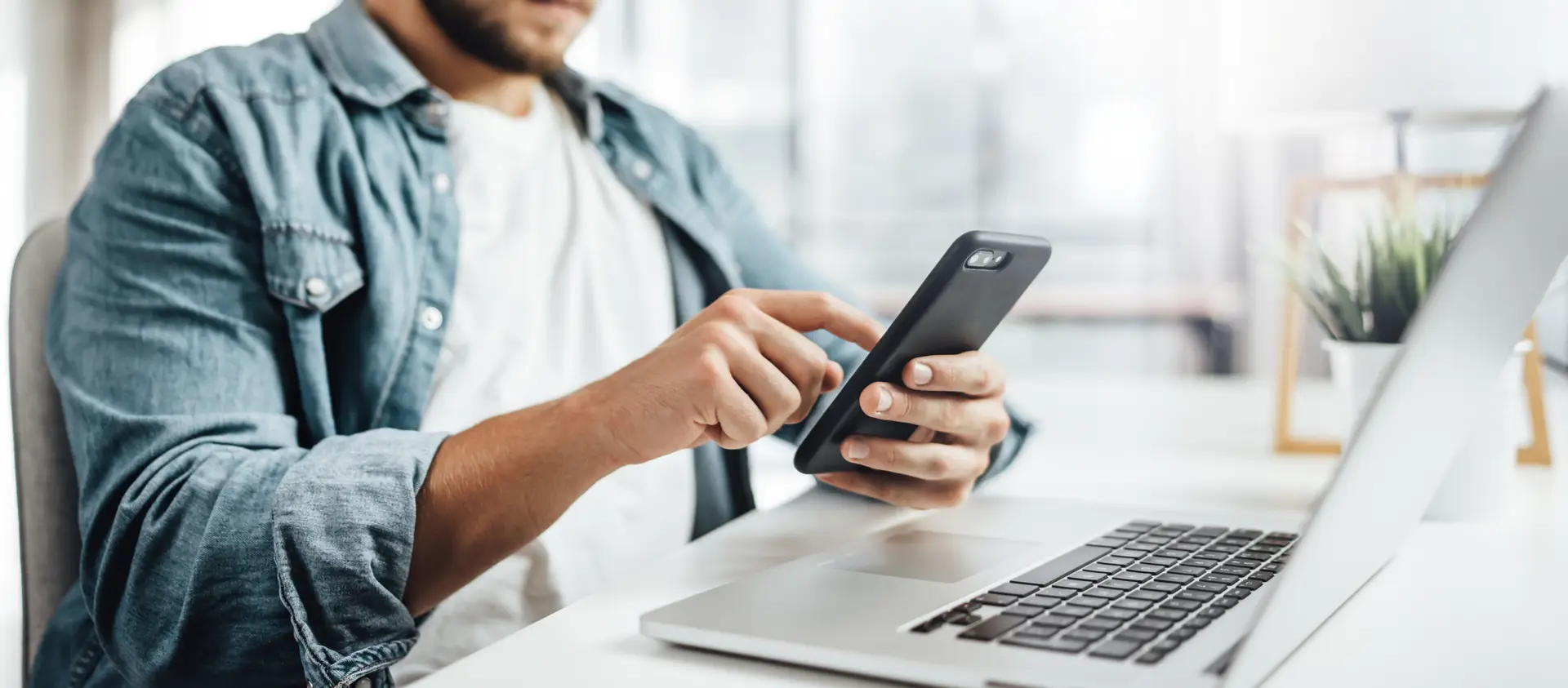 Person using a smartphone while sitting at a desk with an open laptop.