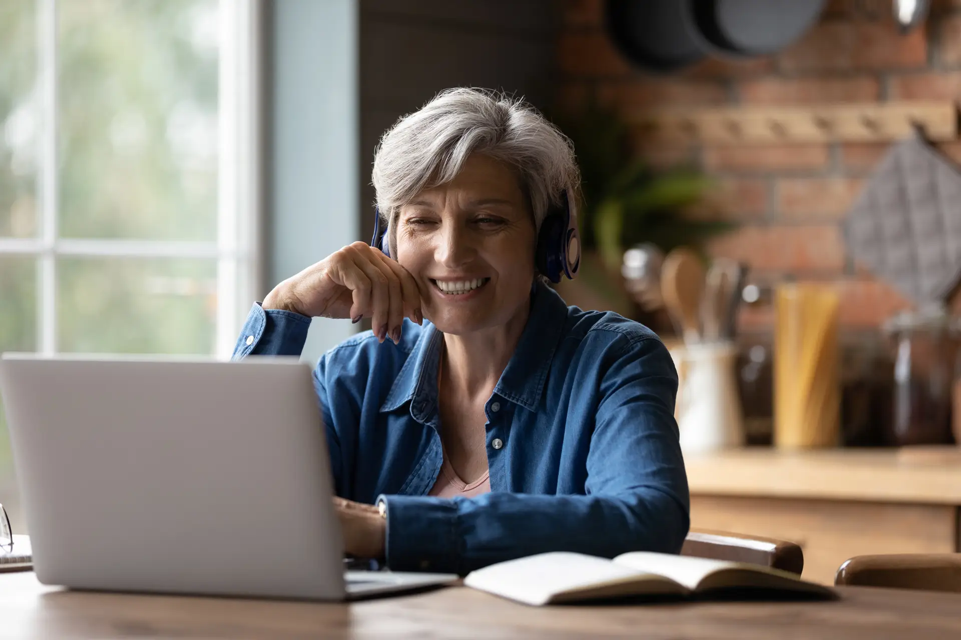 Older woman wearing headphones smiles while using a laptop at a table, participating in a video call or online meeting from home.