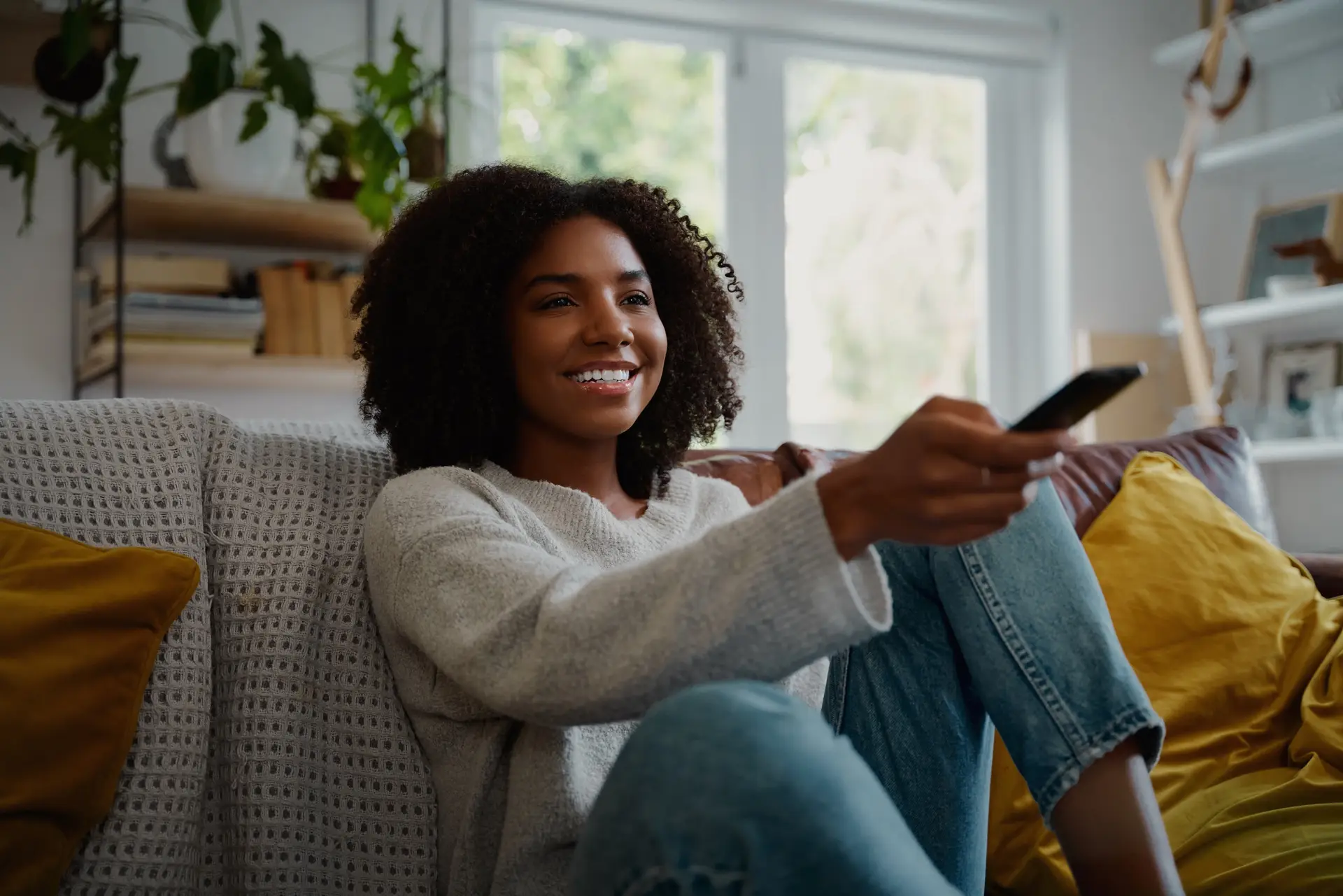 Person sitting on a couch and smiling while holding a TV remote in a bright living room.