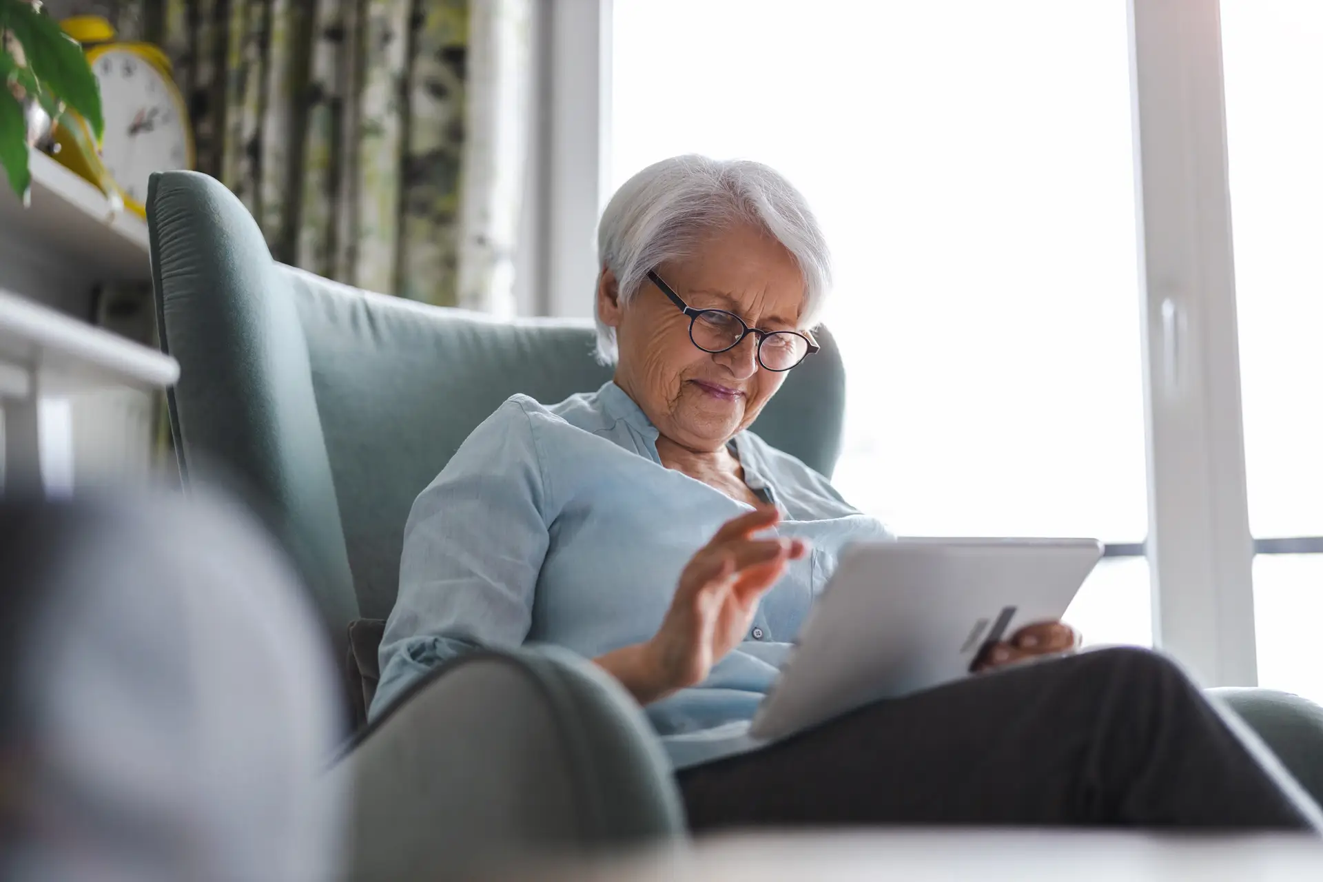 An older woman seated in a cushioned chair taps on a tablet screen while reading, with natural light coming through a nearby window.