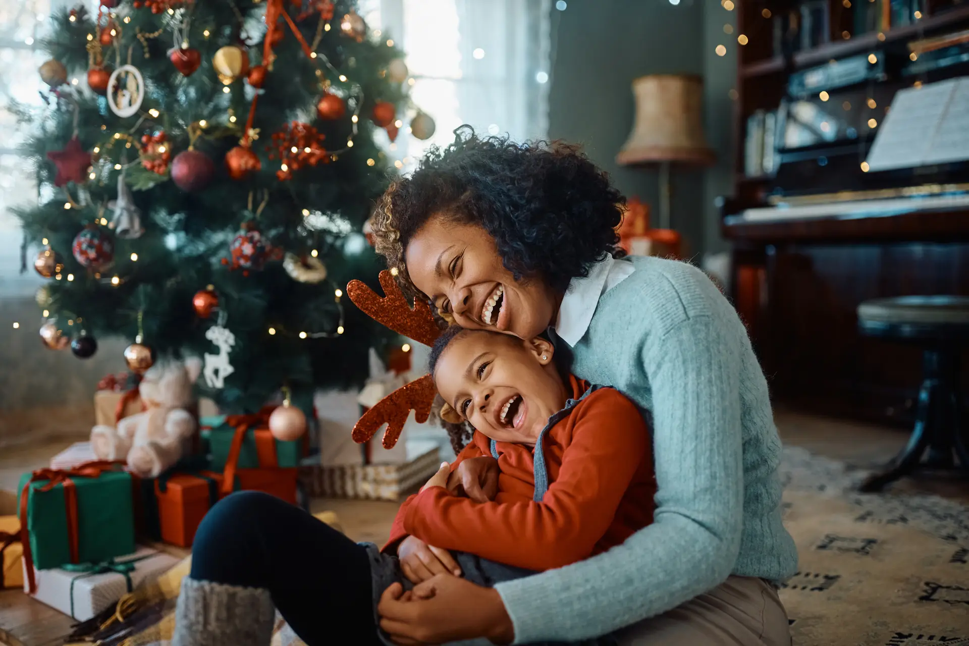 Adult and child laughing and hugging on the floor beside a decorated Christmas tree with wrapped presents and holiday lights.