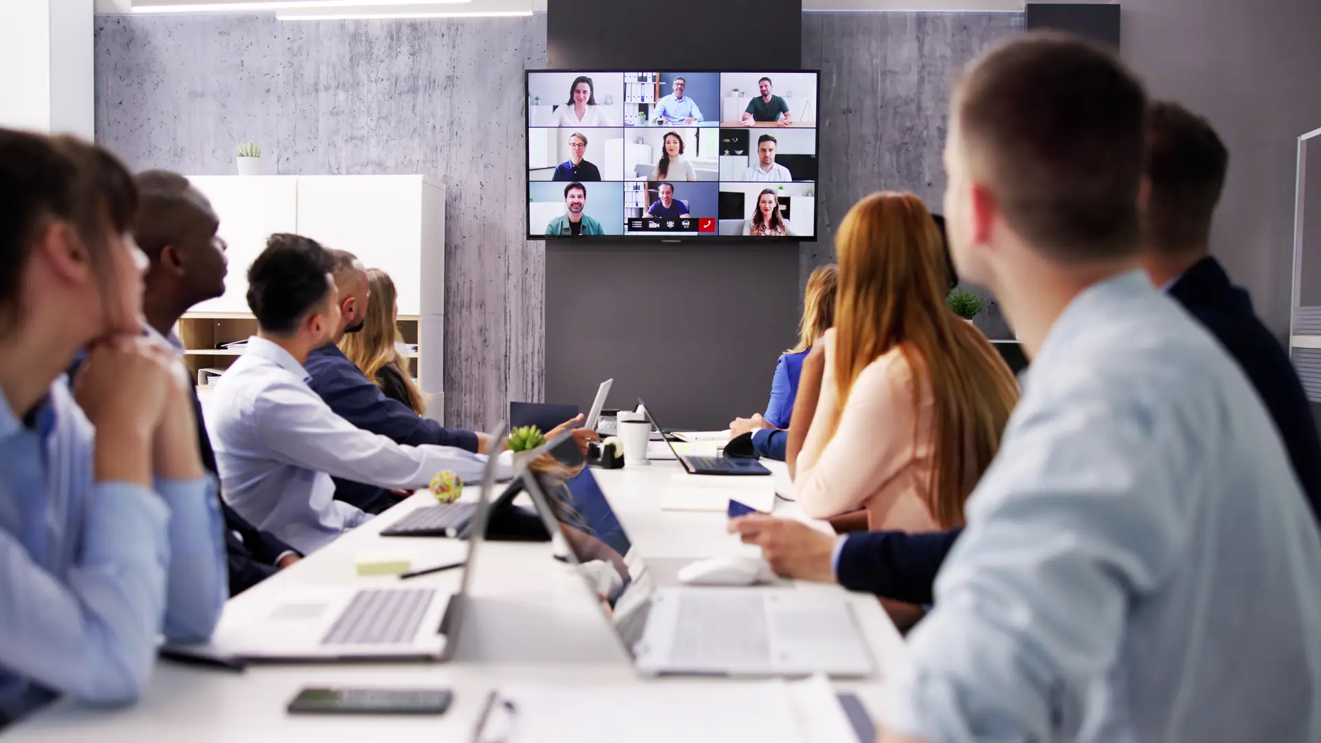 Team members in a conference room participating in a video meeting displayed on a large screen.