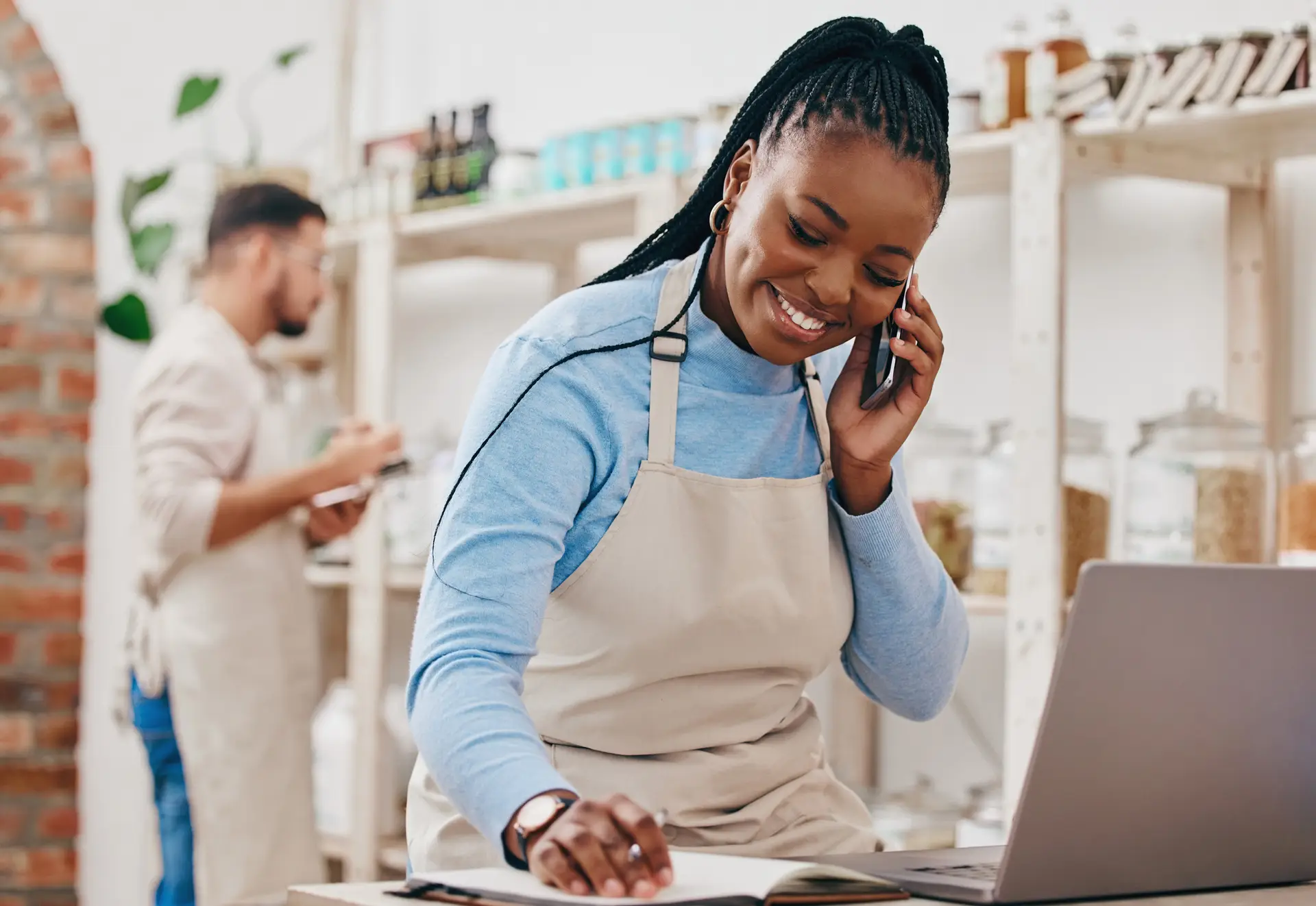 A smiling small-business owner wearing a light blue shirt and beige apron talks on a smartphone while reviewing notes beside an open laptop in a bright shop or café.