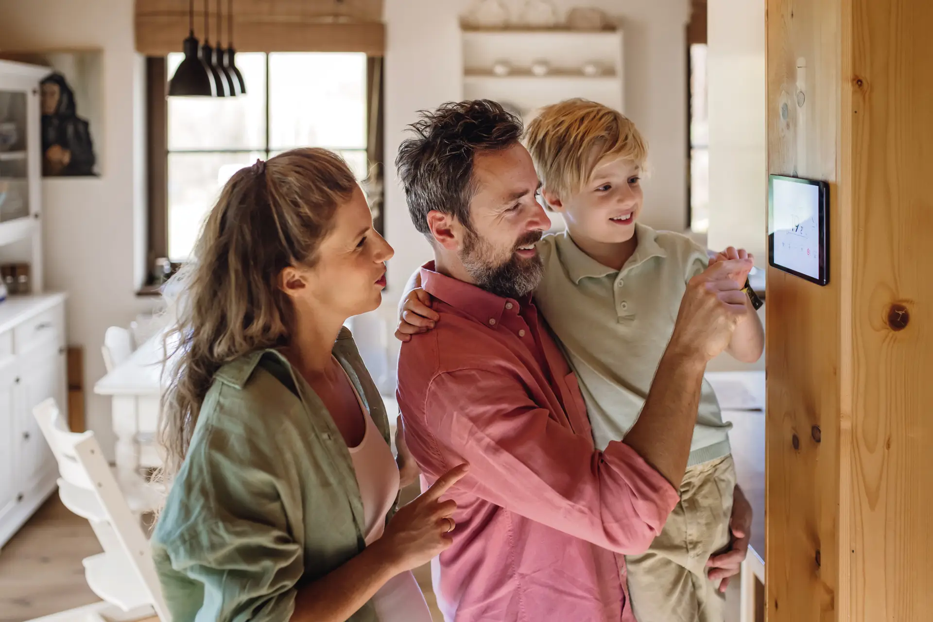 A family gathers around a wall-mounted smart home touchscreen in a bright, modern kitchen. A man holds a young child while they interact with the display, and a woman stands beside them watching with interest.