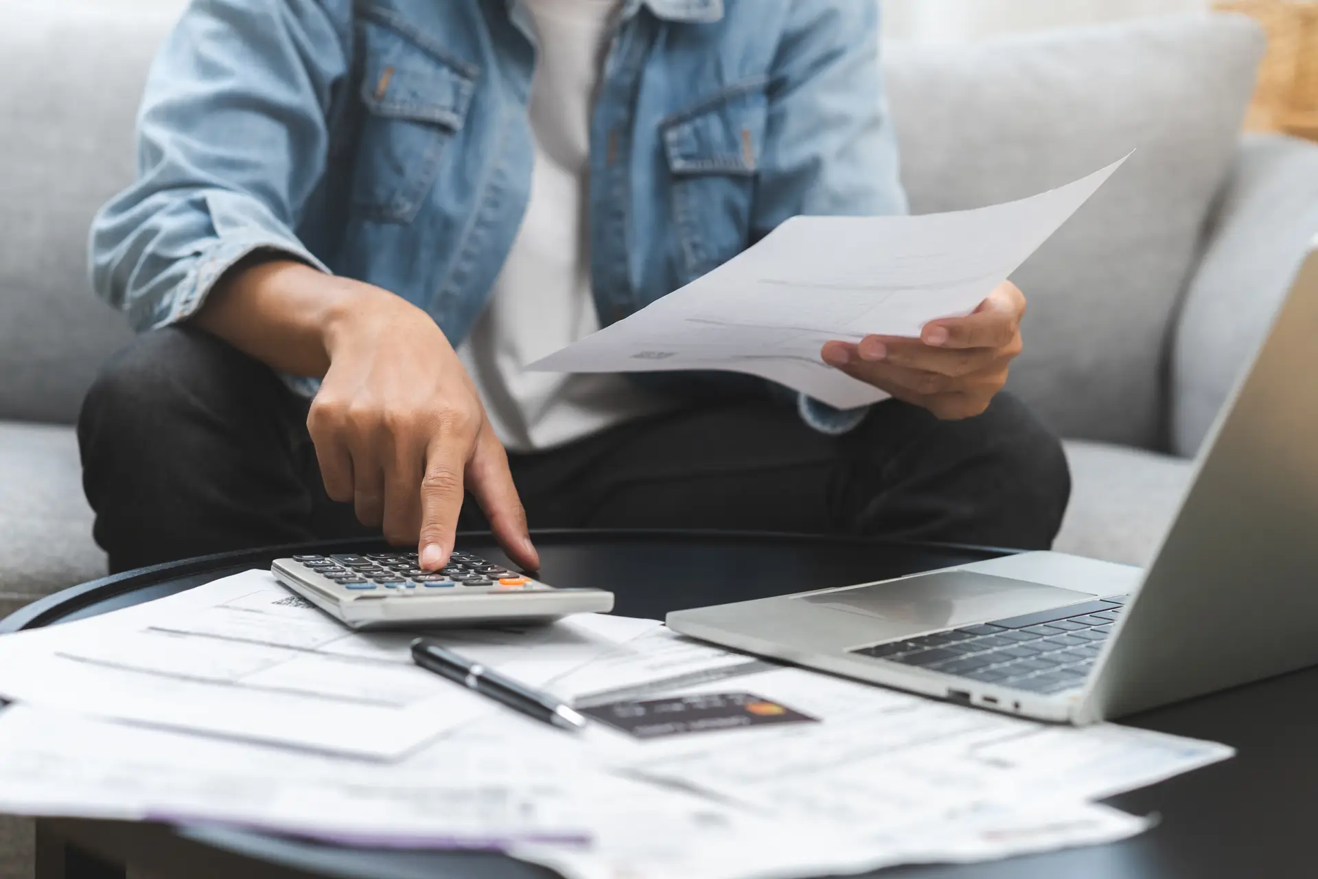 Person reviewing bills and paperwork while using a calculator and laptop at home.