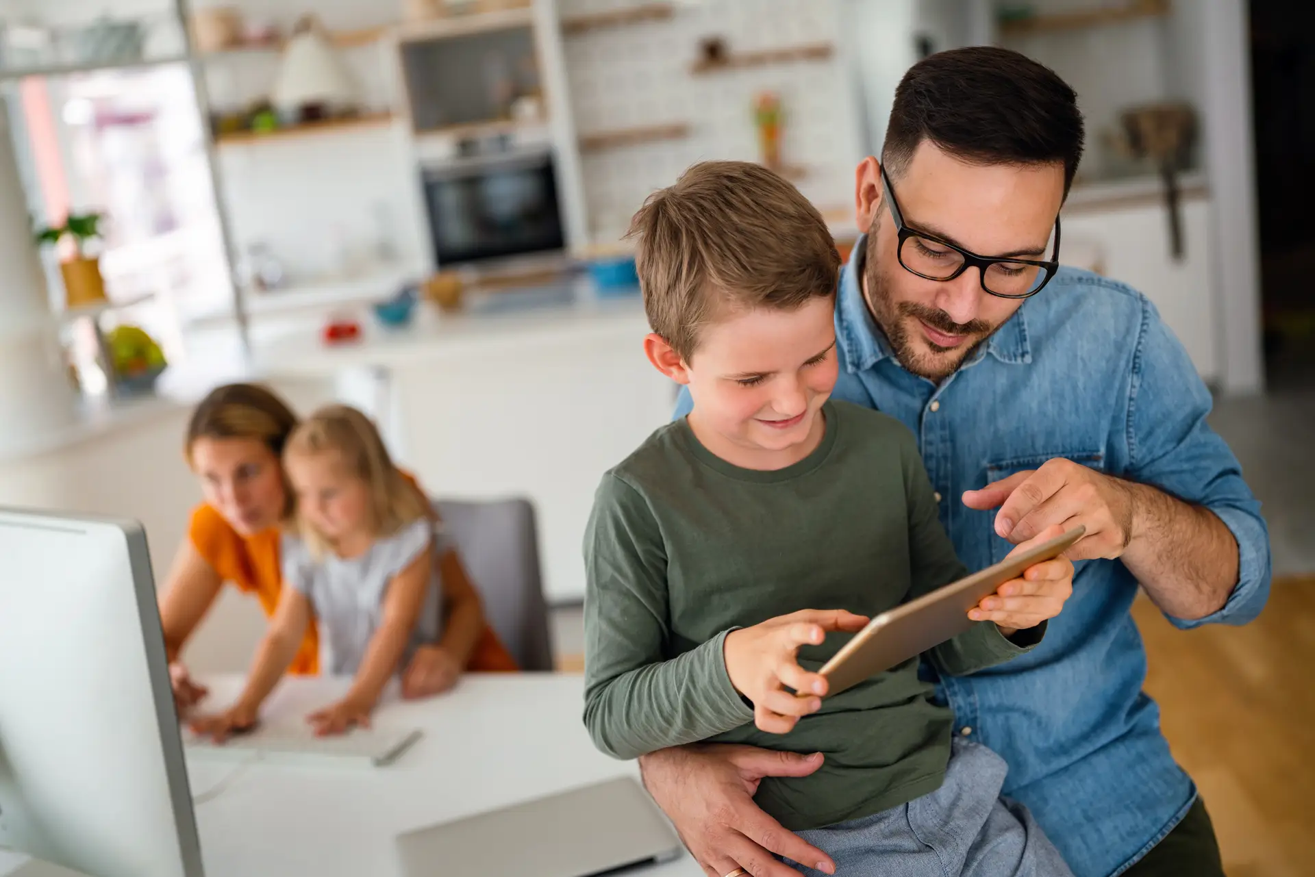 Father and son smiling while using a tablet together at home, with family in the background.