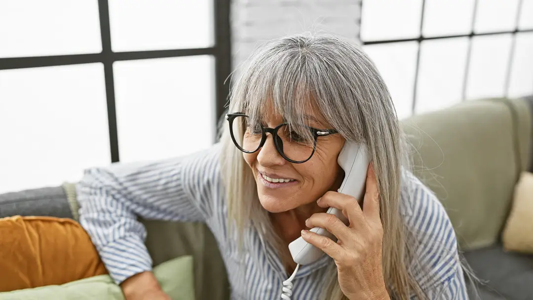 Older woman with gray hair and glasses smiling while talking on a corded telephone in a bright living room.