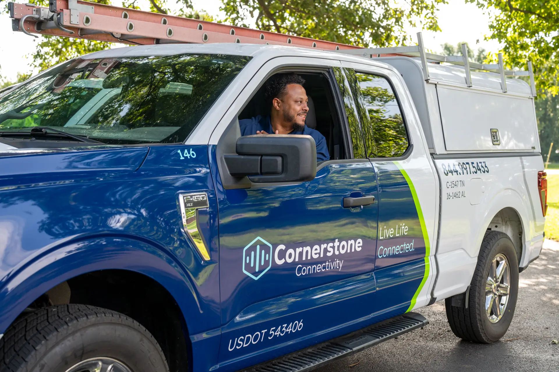 Technician smiles from the driver’s seat of a Cornerstone Connectivity service truck parked outdoors beneath trees.