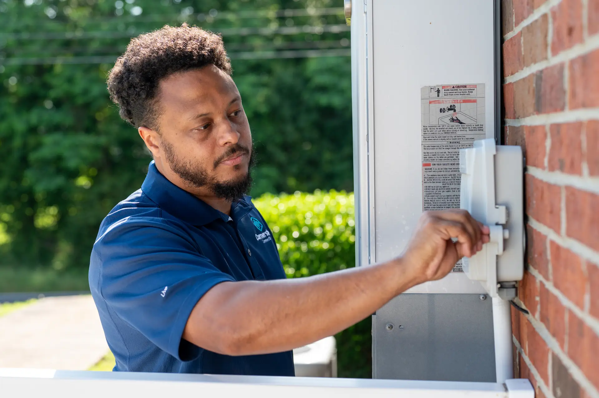 Cornerstone Connectivity technician in a blue work shirt inspecting and servicing an outdoor utility box mounted on a brick wall.