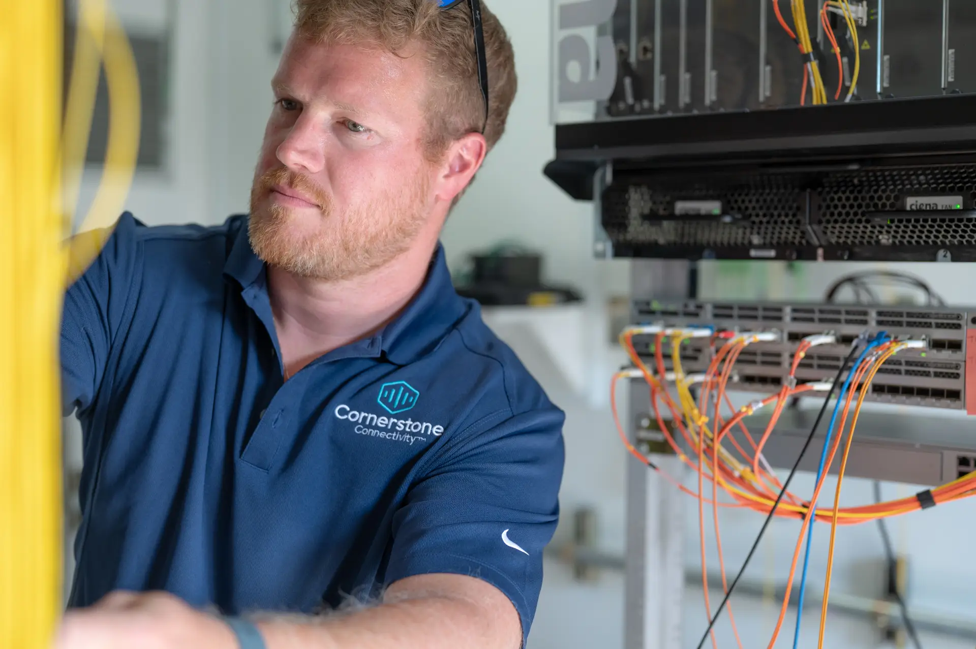 Technician in a blue company polo works on network equipment and fiber optic cables inside a communications rack.