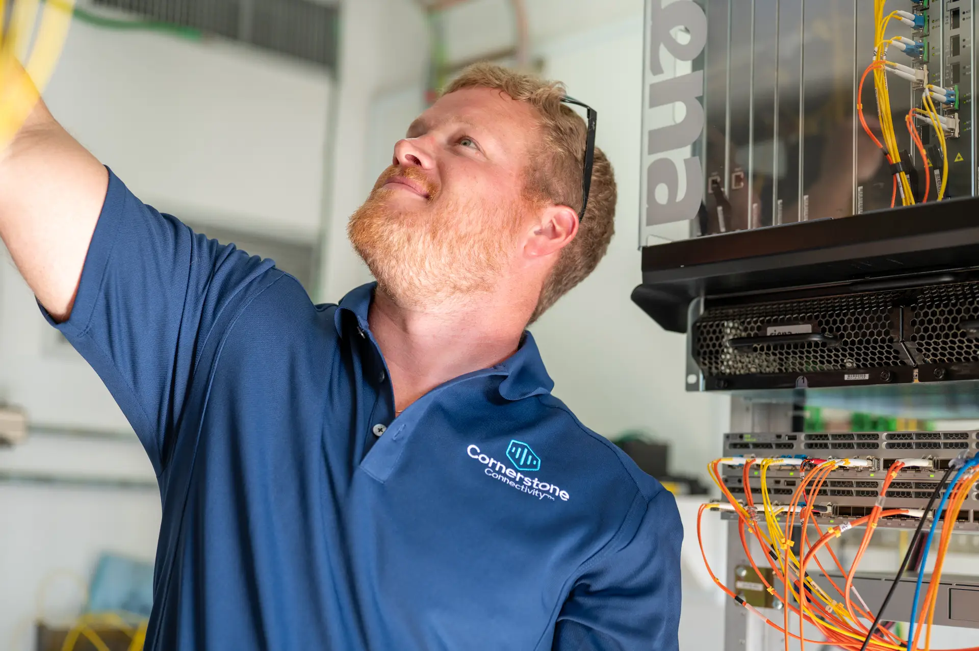 A technician wearing a blue Cornerstone Connectivity polo shirt inspects network cables attached to a server rack.