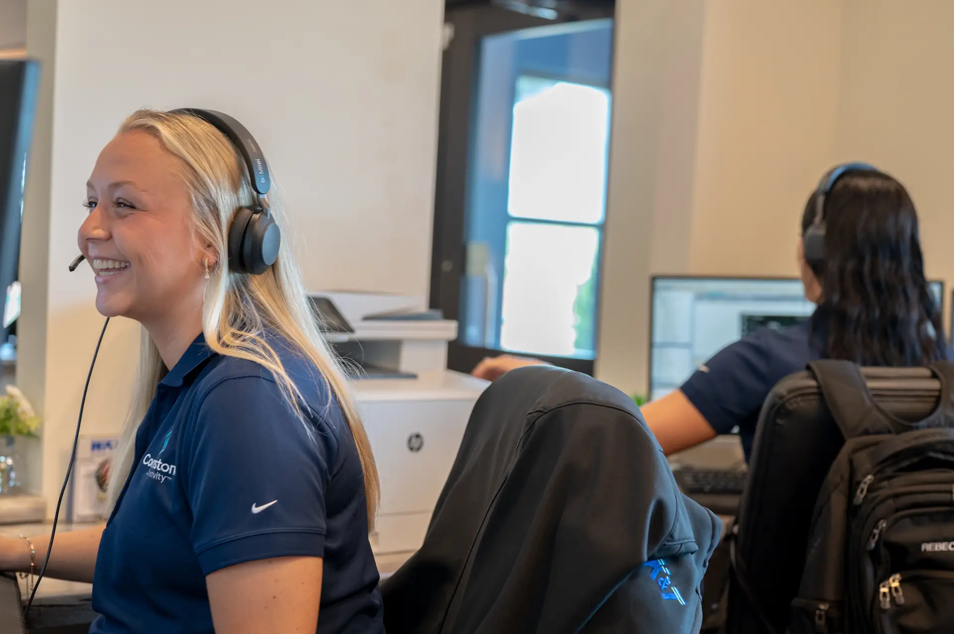 A smiling customer service representative wearing a headset, working at a desk with a computer in a modern office environment.