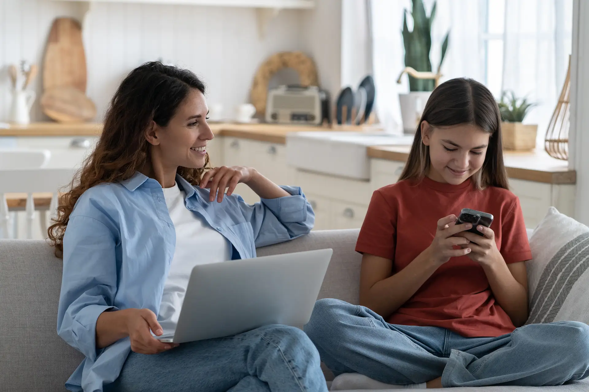 Woman using a laptop and teenage girl using a smartphone while sitting together on a couch at home.