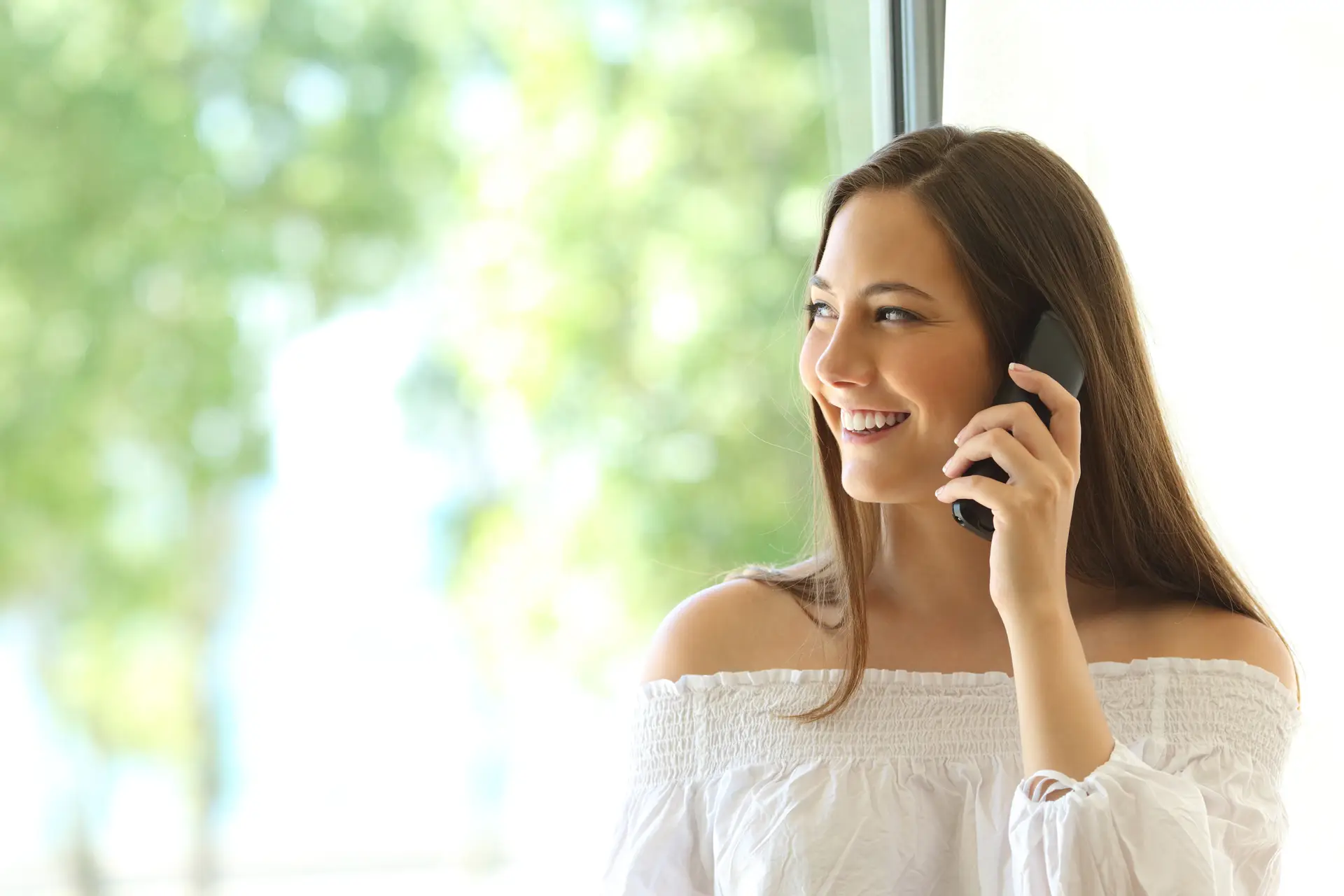 Smiling woman talking on a smartphone near a bright window with greenery visible outside.