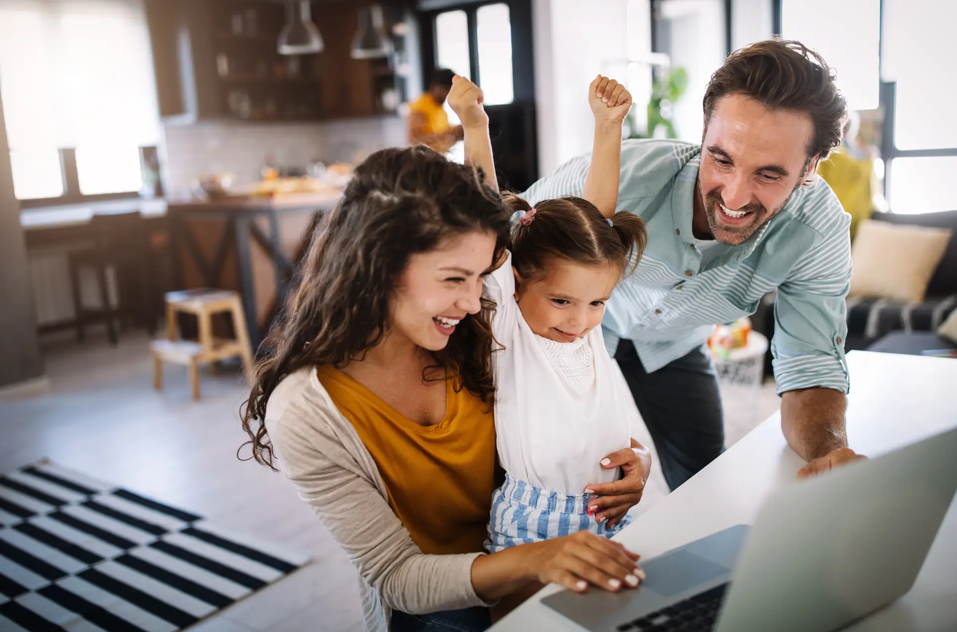 A family gathered around a laptop in a bright home, with two adults smiling and a young child raising her arms excitedly while looking at the screen. The scene conveys a positive shared online experience, with a kitchen and living area visible in the background.