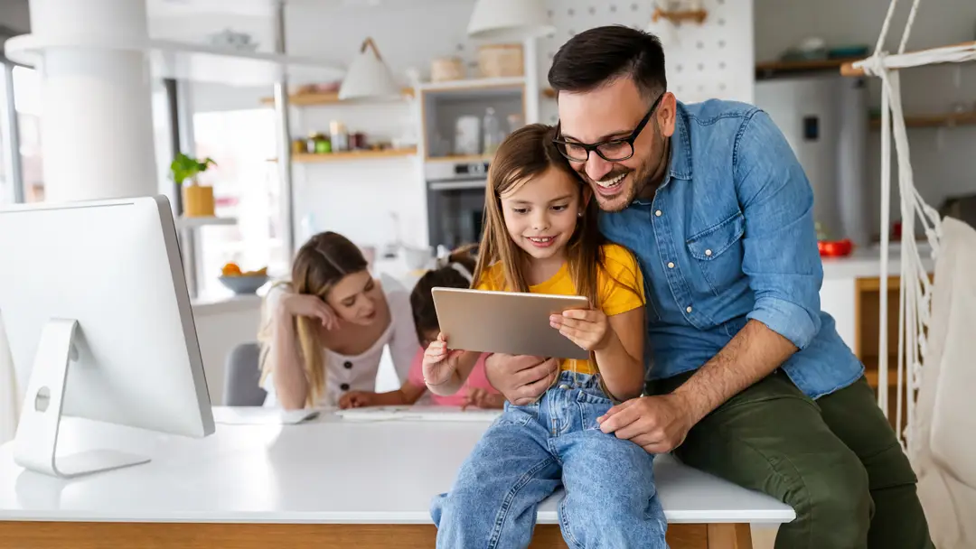 A father and daughter smile while looking at a tablet together in a bright kitchen, with a mother and another child sitting at a desk in the background.
