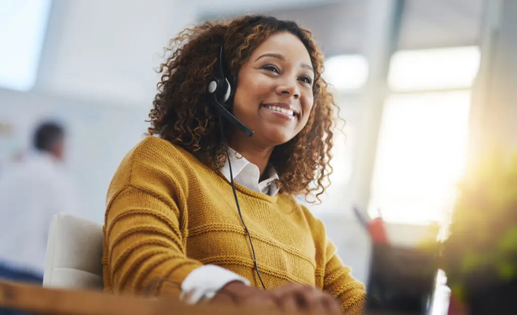 A smiling woman with curly hair wearing a headset while working at her desk in a bright office environment.