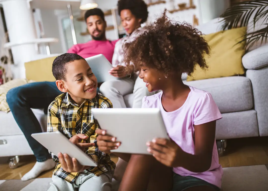 Two children sitting on the floor in a living room, smiling and showing each other tablets. In the background, two adults sit on a sofa using digital devices, creating a family scene centered around technology and connected home activities.