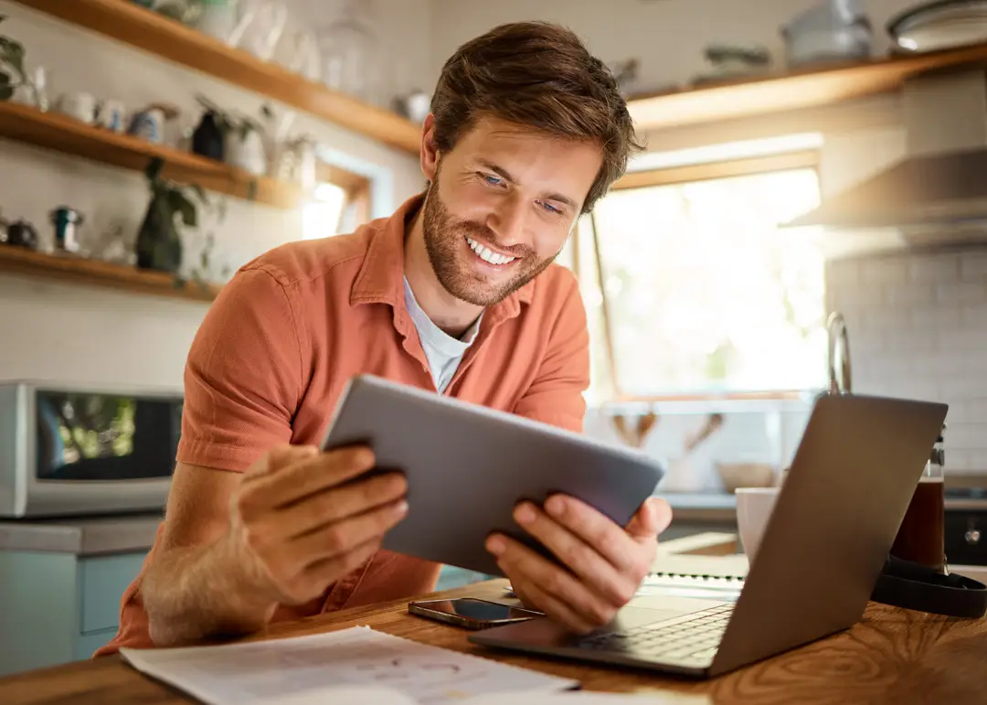 Smiling man standing at a kitchen table, holding a tablet and looking at its screen. An open laptop, smartphone, and papers are on the table in front of him, while shelves, plants, and kitchen items are visible in the bright, sunlit background.