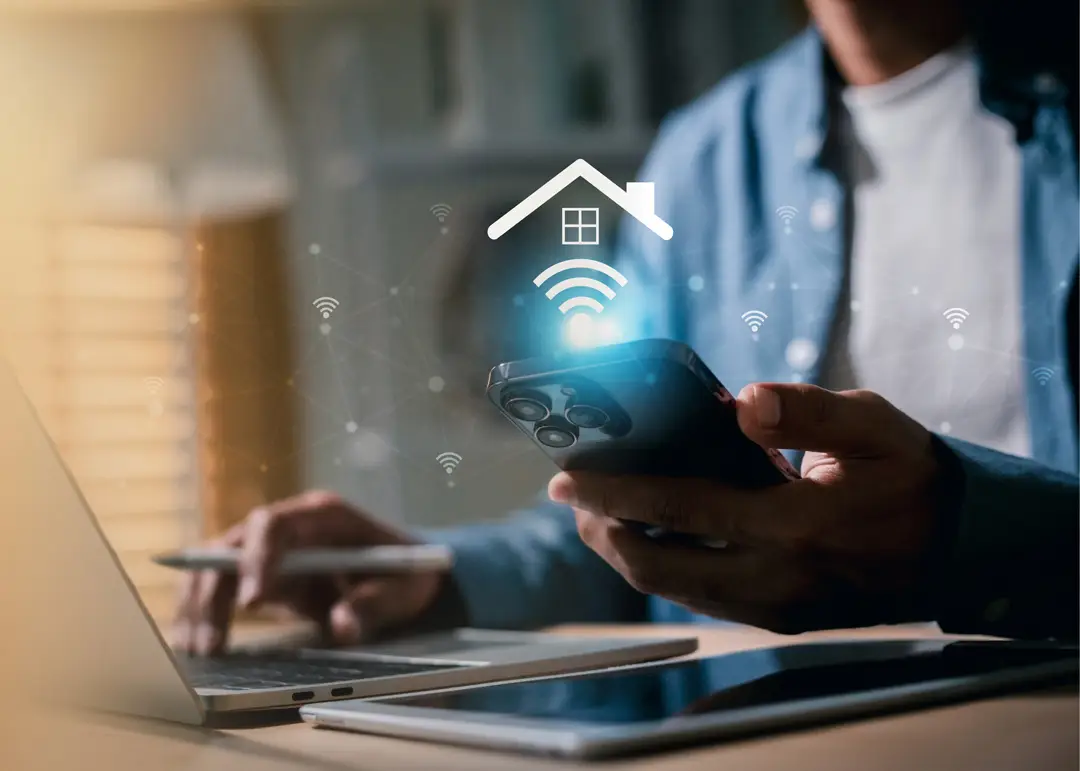 Person using a smartphone while working on a laptop at a desk, with a tablet nearby. A digital smart-home graphic showing a house and wireless connectivity icons appears above the phone, representing remote home automation and connected devices.