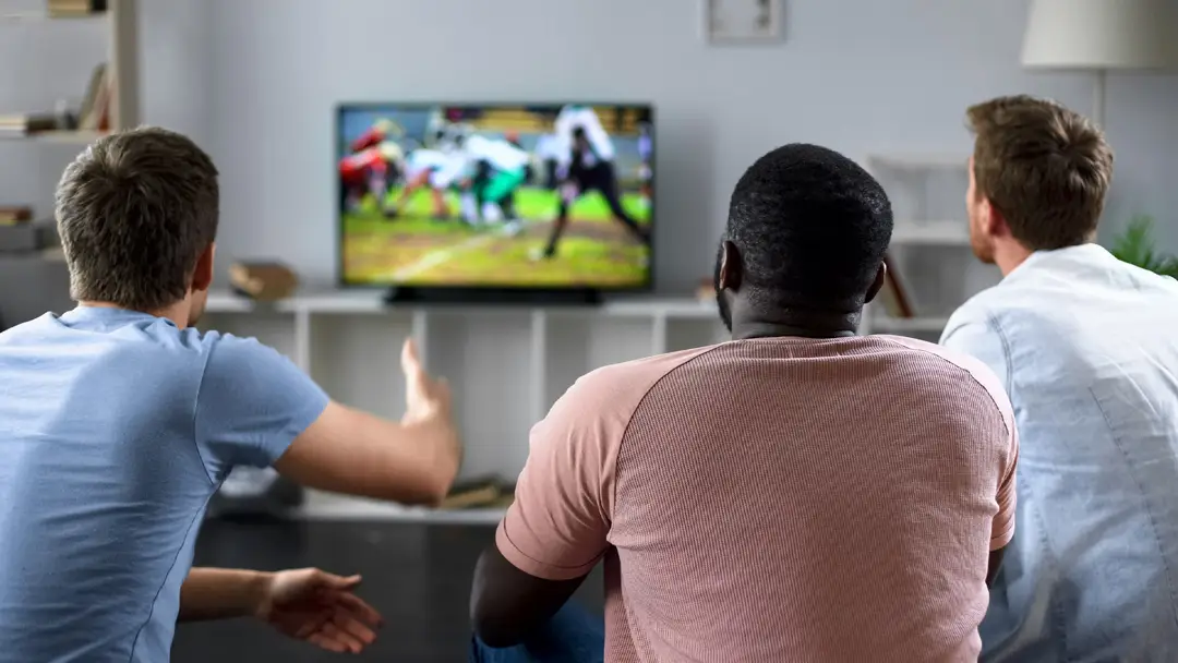 Three male friends seen from behind, sitting in a living room and watching an American football game on a television screen.